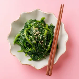 A close-up, top-down shot of a vibrant Korean Spinach Side Dish, showcasing its glistening texture from sesame oil and a sprinkle of toasted sesame seeds in a white bowl.
