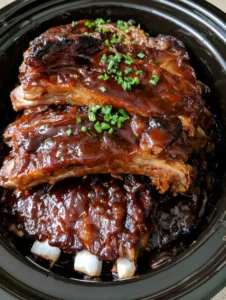 Top-down view of a stack of pork ribs in a black slow cooker with a glossy BBQ sauce. (Crockpot BBQ Ribs)
