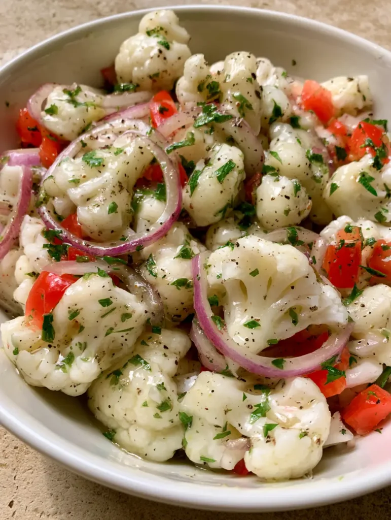 Overhead view of glistening Marinated Cauliflower Salad with red peppers and parsley in a white ceramic bowl.