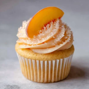 A single gourmet Peach Bellini cupcake with a high peach frosting swirl and vertical fruit slice on a neutral background. (Peach Bellini Cupcakes)