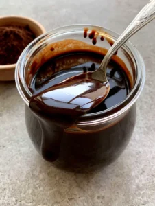 A clear glass jar filled with viscous, reflective Homemade Chocolate Syrup with a silver spoon resting inside.